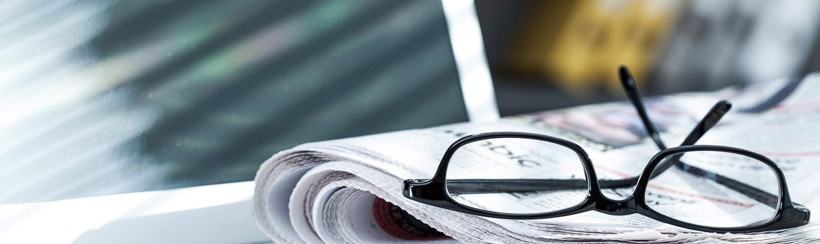 Eine Brille liegt auf einer aufgeschlagenen Zeitung, während Sonnenlicht durch ein Fenster fällt.