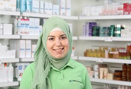 Eine lächelnde Frau in einem grünen Kopftuch steht in einer Apotheke vor Regalen mit Medikamenten.