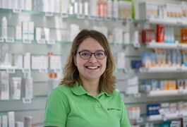 Eine lächelnde Frau im grünen Polo steht in einer Apotheke vor Regalen voller Gesundheitsprodukte.