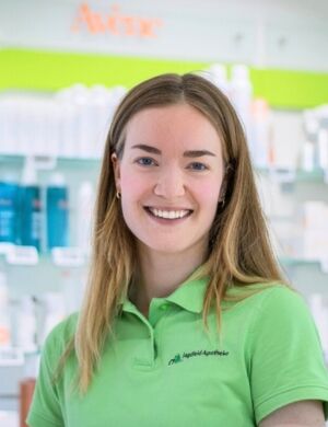Eine Frau in grüner Arbeitskleidung lächelt freundlich in einer Apotheke als Ansprechpartnerin.
