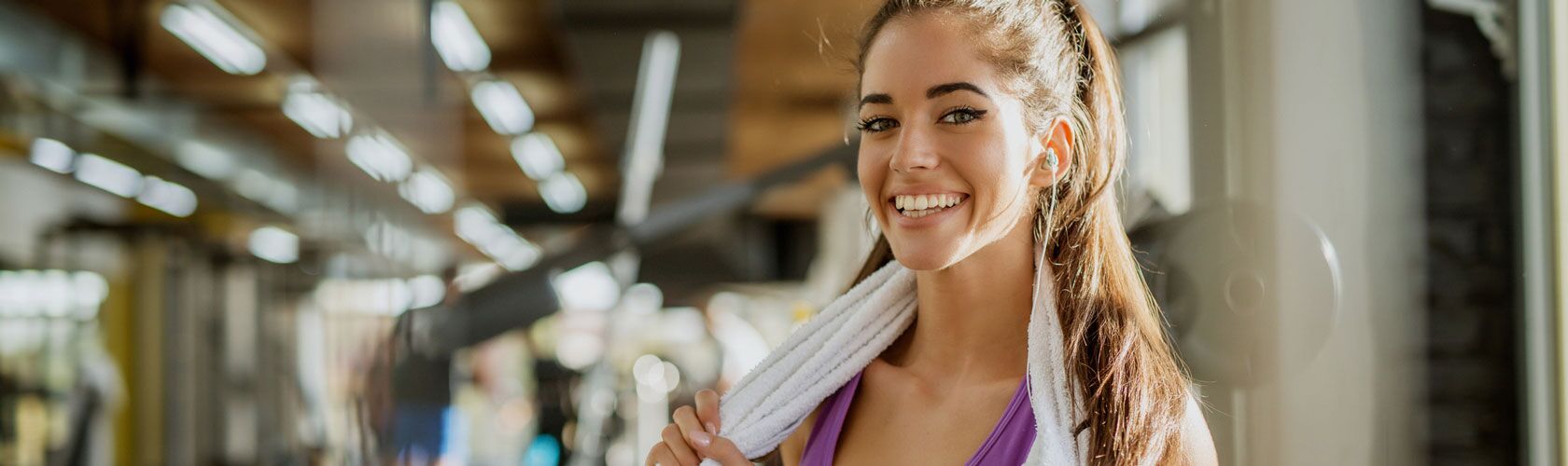 Eine lächelnde Frau in Sportkleidung steht mit einem Handtuch im Fitnessstudio, was auf die Vorteile von Fitness hinweist.